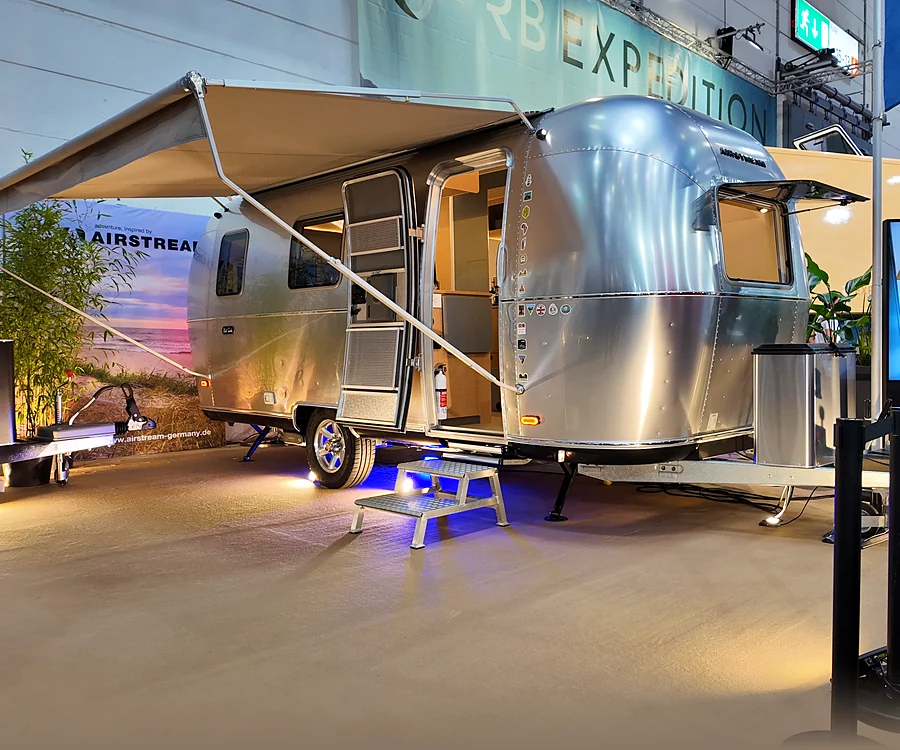 Wohnwagen mit Aluminiumaufbau und ausgefahrener Markise auf einem Messestand