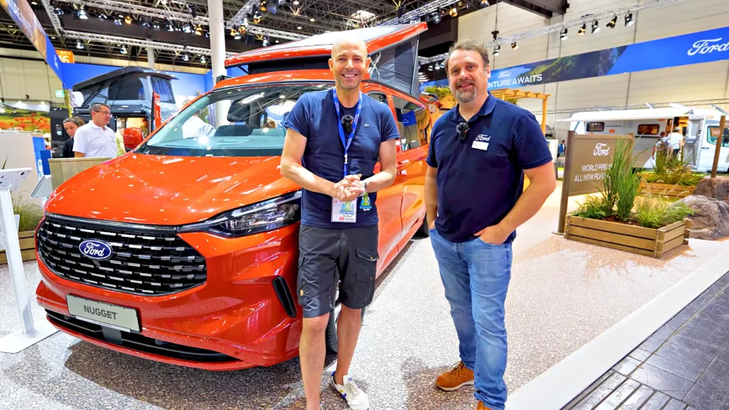 Thomas von CampingTurtle und Tiemo Etzold stehen vor dem orangefarbenen Ford Nugget mit aufgestelltem Dach auf dem Caravan Salon 2023.