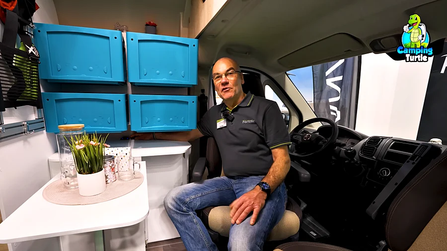 Detlef sitzt auf dem gedrehten Fahrersitz vor dem kleinen Tisch, darüber vier blaue Boxen