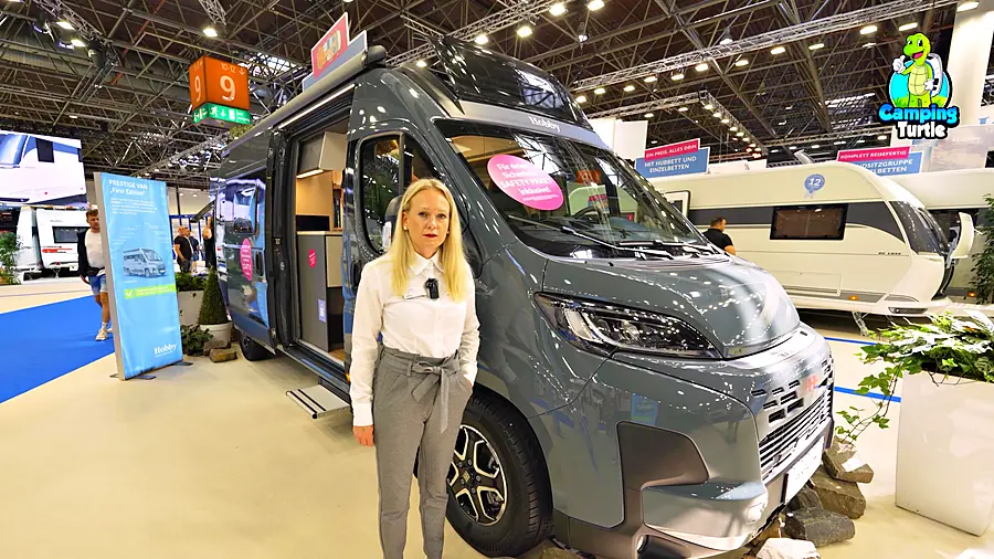 Blonde Frau in weißer Bluse steht allein vor dem rechten Scheinwerfer eines blaugrauen Kastenwagens auf einem Messestand