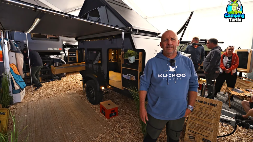 Peter Schneewolf steht vor dem grauen Mini-Wohnwagen mit Dachzelt, Markise und offener Tür