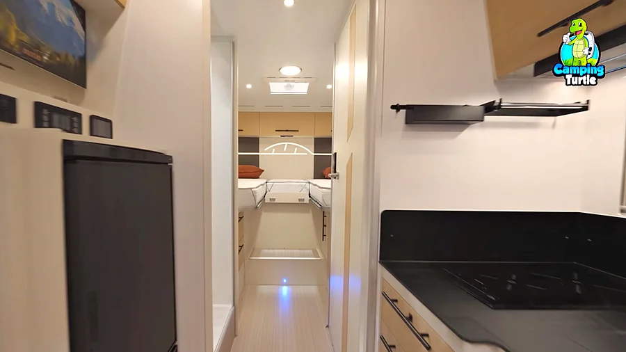 Blick durch den Mittelgang mit dunklem Kühlschrank und Bedienpanels links, Küchenzeile mit schwarzer Arbeitsplatte rechts und den beiden Einzelbetten im Heck