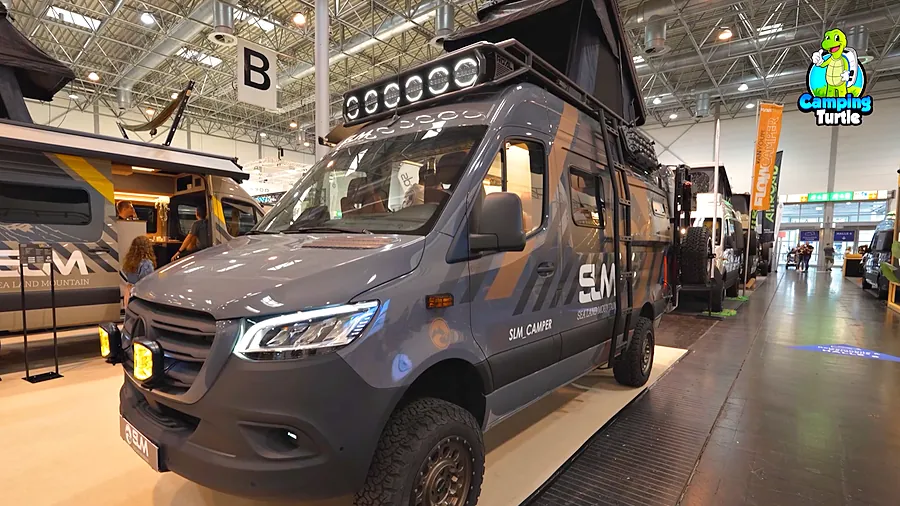 Grauer SLM SPR 600 auf Mercedes Sprinter schräg von vorn mit Dachträger und LED-Lichtleiste, Leiter am Heck, grobstolligen Reifen und gelben Zusatzscheinwerfern