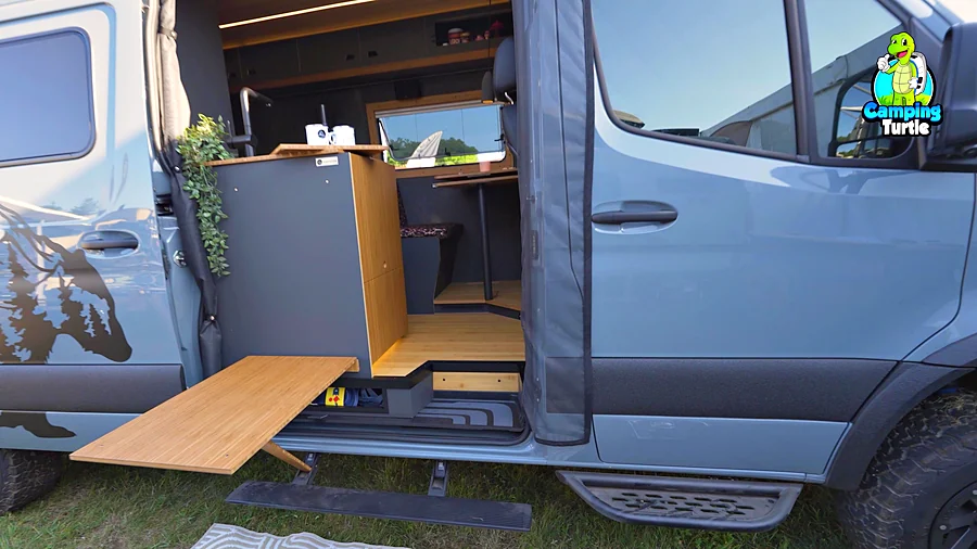 Holzplatte, die waagerecht aus der offenen Schiebetür eines grauen Vans ragt und von einem schrägen Tischbein auf der Trittstufe gestützt wird, dahinter ein grauer Küchenblock mit Bambusseite und ein heller Holzboden
