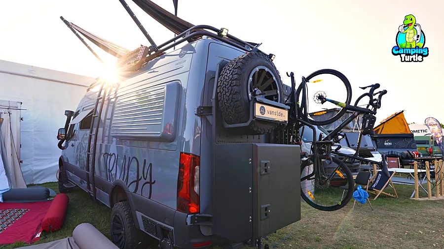 Heck eines grauen Vans mit Reserverad, darunter eine dunkle Staubox, daneben zwei Fahrräder am Fahrradlift, darüber die Sonne