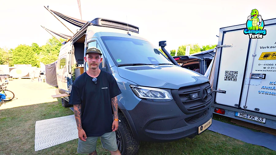 Mann mit Kappe und schwarzem T-Shirt vor einem grauen Mercedes Sprinter mit Dachträger und Schnorchel auf einer Wiese