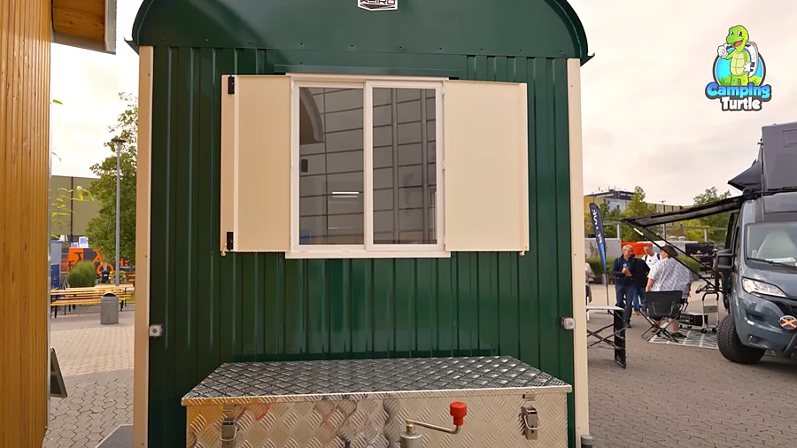 Front des grünen Rolling Home mit gewölbtem Dach, einem großen zweiteiligen Fenster mit zwei aufgeklappten cremefarbenen Fensterläden und einer Box aus Riffelblech auf der Deichsel