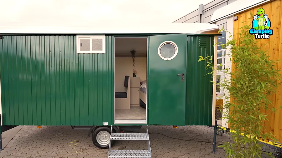 Grüner Rolling Home 470 von der Seite mit geöffneter Stahltür mit Bullauge, davor eine eingehängte Treppe mit Gitterroststufen und durch die Tür der Blick auf Sitzbank, kleinen Schrank und Bett