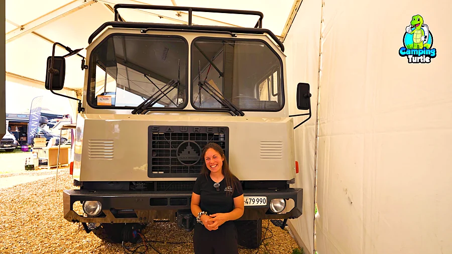 Junge Frau in schwarzem T-Shirt steht lächelnd vor der Front eines hellgrauen Lkw mit Saurer-Kühlergrill und Dachgepäckträger in einem Messezelt
