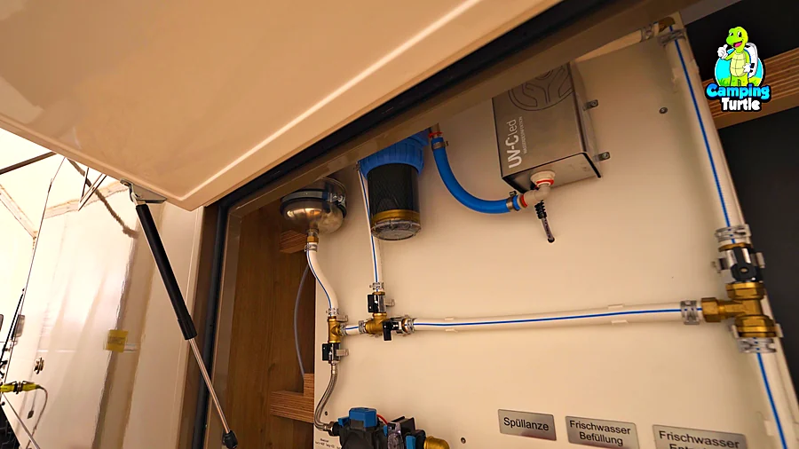 Geöffnete Klappe an einer hellen Kabinenwand mit blauen Wasserleitungen, Filtergehäuse, UV-Einheit und beschrifteten Absperrhähnen